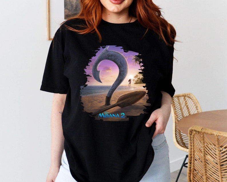 Puede incluir: Camiseta negra con un estampado gr&aacute;fico de un gancho azul y una pala de madera en una playa con una puesta de sol de fondo. El texto "Moana 2" est&aacute; impreso debajo del gr&aacute;fico.