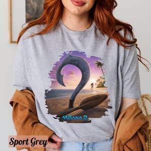 Puede incluir: Una camiseta gris con un gr&aacute;fico de un gancho de dibujos animados y un barco en una playa. El texto "Moana 2" est&aacute; impreso en la camiseta.