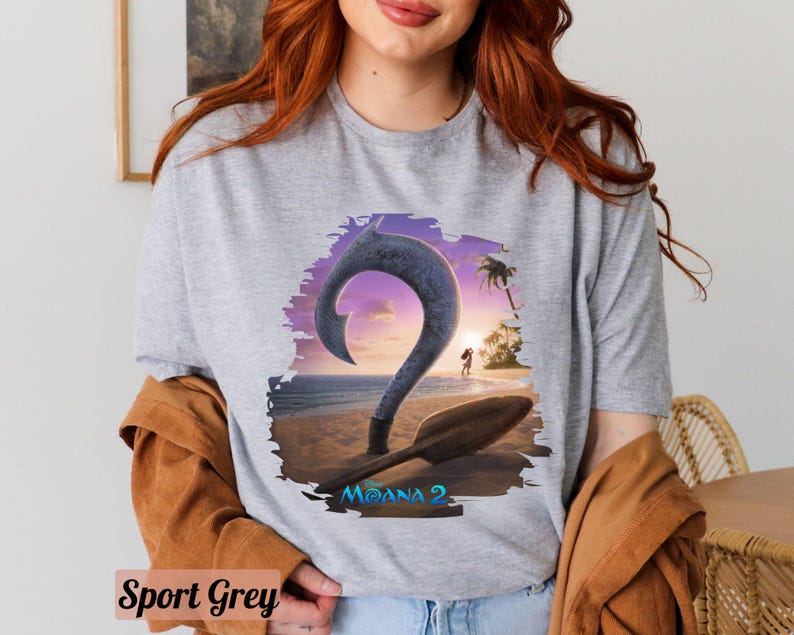 Puede incluir: Una camiseta gris con un gr&aacute;fico de un gancho de dibujos animados y un barco en una playa. El texto "Moana 2" est&aacute; impreso en la camiseta.