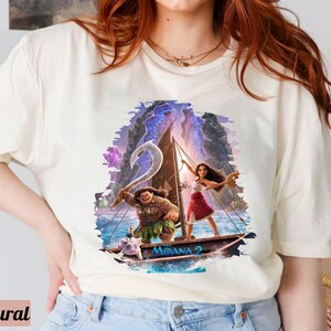 May include: A white t-shirt featuring a colourful graphic of Moana and Maui sailing on a boat. The graphic includes the text "Moana 2".