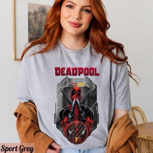 May include: A gray t-shirt with a graphic of Deadpool from Marvel Studios. The graphic shows Deadpool in a red and black suit with his hands up in a fighting pose. The text "DEADPOOL" is printed in red above the graphic.