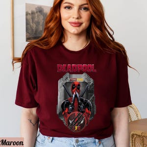 May include: A maroon t-shirt with a graphic of Deadpool from Marvel Studios. The graphic shows Deadpool with his hands over his mouth and a red and black mask. The text "DEADPOOL" is above the graphic and "MARVEL STUDIOS" is below the graphic.