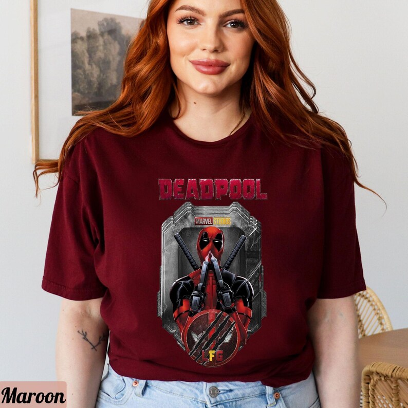 May include: A maroon t-shirt with a graphic of Deadpool from Marvel Studios. The graphic shows Deadpool with his hands over his mouth and a red and black mask. The text "DEADPOOL" is above the graphic and "MARVEL STUDIOS" is below the graphic.