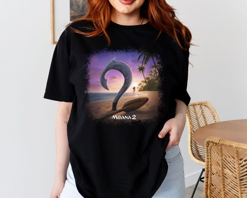 May include: Black t-shirt with a graphic print of a sunset scene with a hook and a paddle. The text "Moana 2" is printed below the graphic.