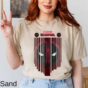 Op de afbeelding: Een beige t-shirt met een rode en zwarte afbeelding van Deadpools gezicht met de tekst "MARVEL STUDIOS DEADPOOL" boven de afbeelding.