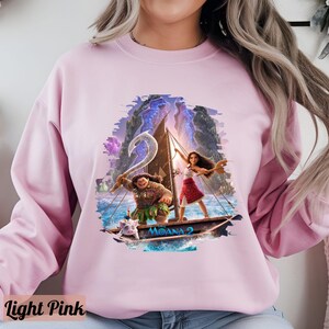 May include: A light pink sweatshirt featuring a graphic print of Moana and Maui sailing on a canoe. The print depicts a blue ocean, green islands, and a pink sky. The text "Moana" is printed on the canoe.