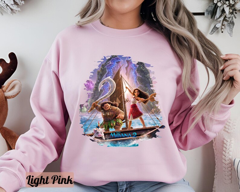 May include: A light pink sweatshirt featuring a graphic print of Moana and Maui sailing on a canoe. The print depicts a blue ocean, green islands, and a pink sky. The text "Moana" is printed on the canoe.