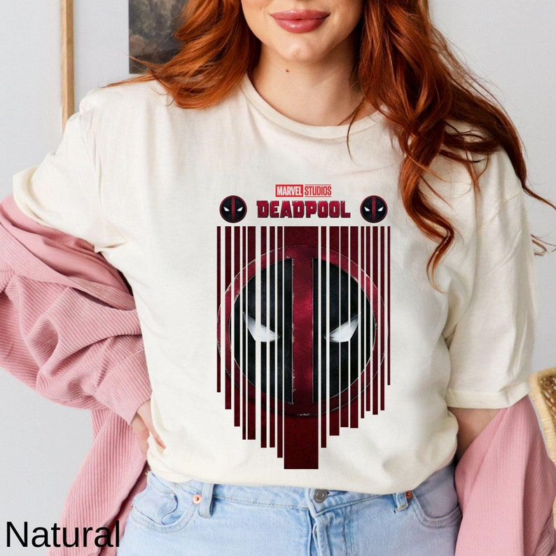 Op de afbeelding: Een wit T-shirt met een rode en zwarte Deadpool-afbeelding. De afbeelding heeft een barcode-ontwerp met het Marvel Studios-logo en het woord "Deadpool" boven de barcode.