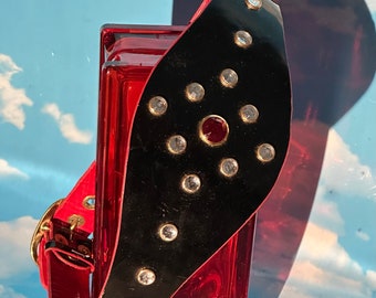 May include: A black leather collar with a red jewel and clear rhinestones. The collar is on a red glass surface.