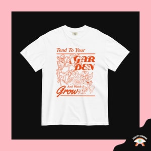 Op de afbeelding: Wit T-shirt met een rood grafisch ontwerp met de tekst "Tend To Your Garden And Watch It Grow". De grafiek toont een gestileerde bloem met een fee.