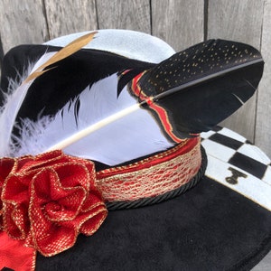 May include: A black and white hat with a red and gold ribbon and a white feather with black tips. The hat has a checkerboard pattern on the brim.