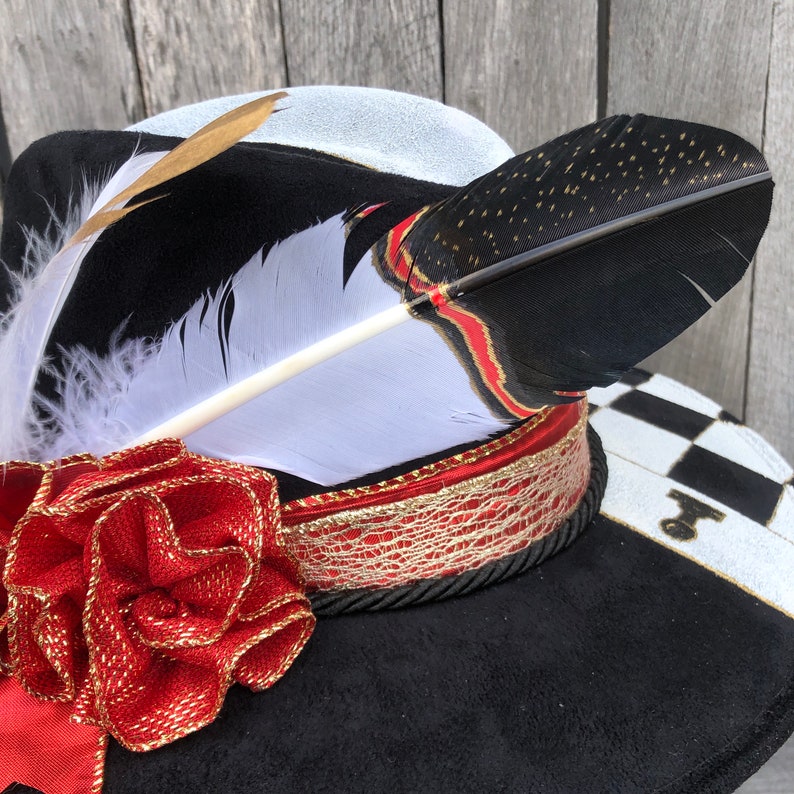 May include: A black and white hat with a red and gold ribbon and a white feather with black tips. The hat has a checkerboard pattern on the brim.