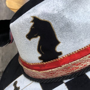 May include: A black and white chess piece in the shape of a knight is painted on the top of a black and white checkered hat. The hat has a red and gold ribbon around the brim.