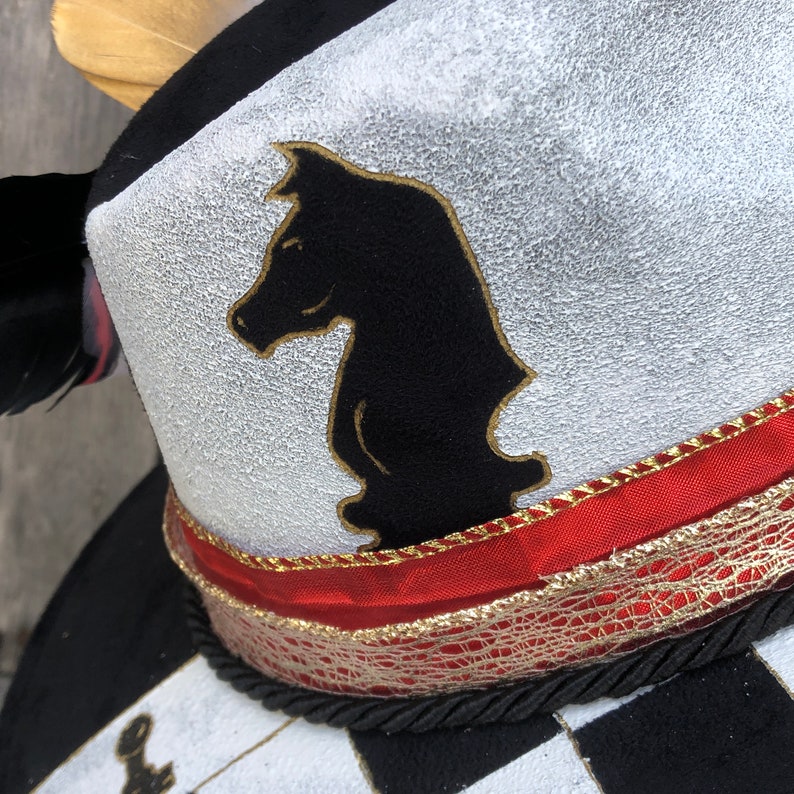 May include: A black and white chess piece in the shape of a knight is painted on the top of a black and white checkered hat. The hat has a red and gold ribbon around the brim.