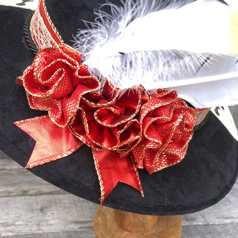 May include: A black velvet hat with a red and gold ribbon and a white feather. The hat is decorated with a large red flower.