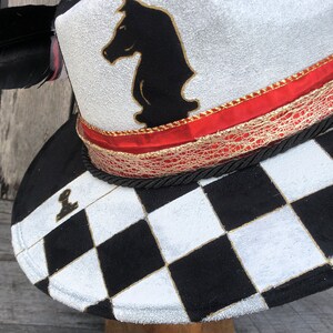 May include: A white hat with a black chess knight design, red and gold trim, and a black and white checkered pattern on the brim.