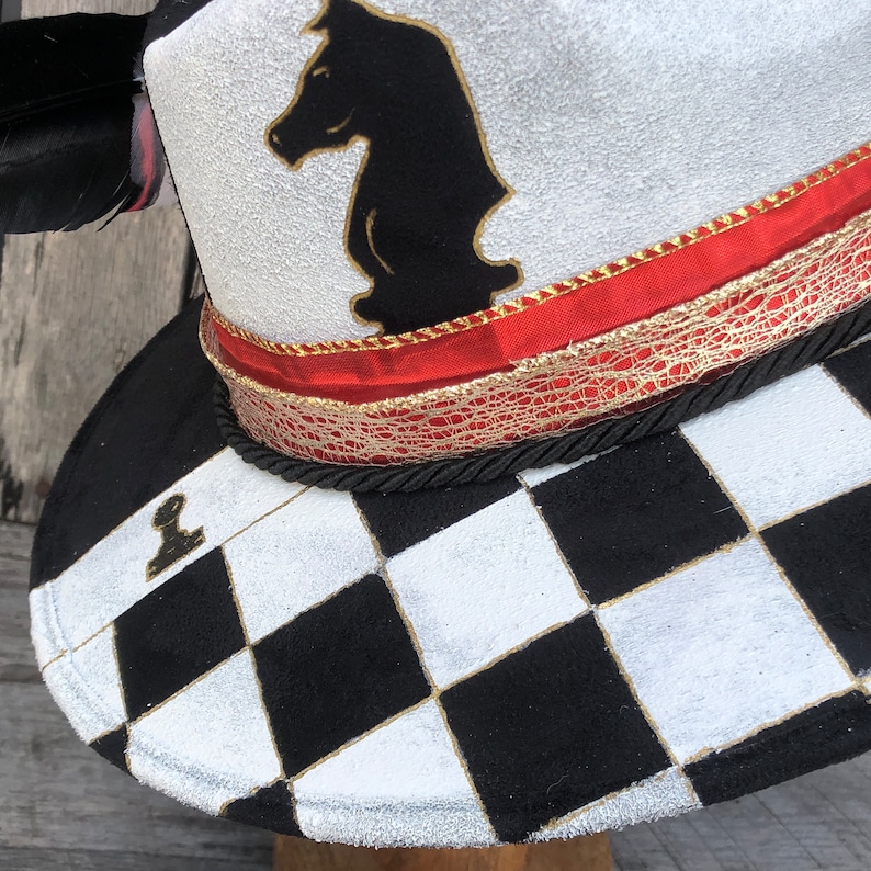 May include: A white hat with a black chess knight design, red and gold trim, and a black and white checkered pattern on the brim.