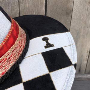 May include: A black and white checkered hat with a gold trim and a red ribbon. The hat has a black chess piece on the top.