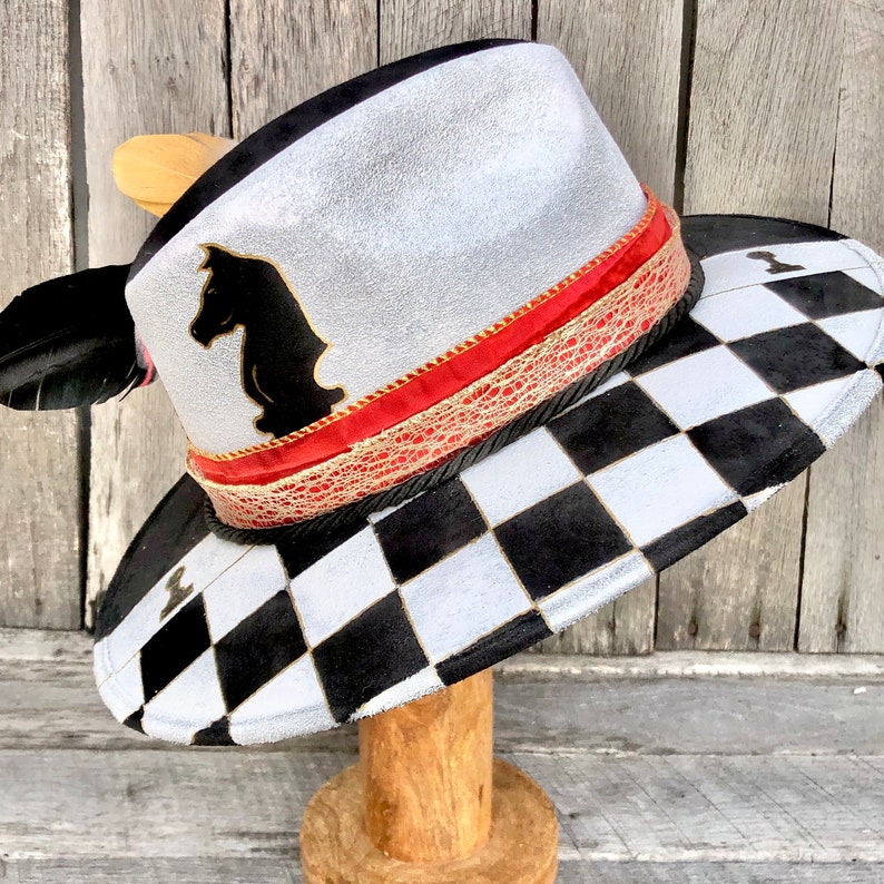 May include: A white and black checkered cowboy hat with a red and gold band. The hat has a black knight silhouette on the front.
