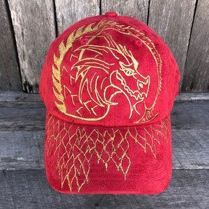 以下が含まれることがあります： 赤いベースボールキャップ。前面に金の刺繍で龍のデザインが施され、つばには金の刺繍模様が施されています。