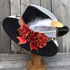May include: A black wide-brimmed hat with a white and gold trim. The hat is decorated with a large red flower and a white feather with black tips.