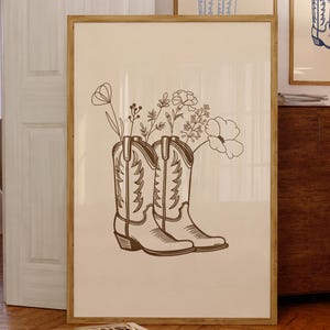Puede incluir: Una impresión enmarcada con un dibujo lineal de dos botas de vaquero con flores que crecen en la parte superior. Las botas son marrones y las flores son blancas y marrón claro.