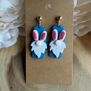 May include: A pair of blue gnome earrings with white beards, pink and white bunny ears, and gold-coloured studs. The earrings are displayed on a brown card.