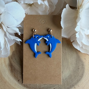 May include: A pair of blue dolphin earrings with white accents, displayed on a brown card. The earrings have gold-colored posts. The background includes white flowers and a wooden surface.