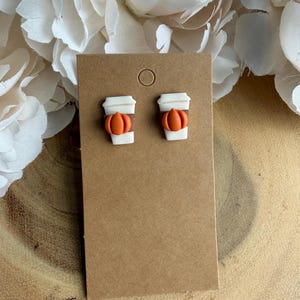 Op de afbeelding: Een paar oorknopjes in de vorm van witte koffiebekers met bruine sleeves en kleine oranje pompoenen aan de voorzijde, gepresenteerd op een bruine kartonnen kaart met een rond gat aan de bovenkant om op te hangen.
