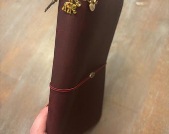 以下が含まれることがあります： 金色のハトメと装飾的なチャームが付いた、バーガンディ色の革製ジャーナル。チャームには、象、ハート、小さな装飾品が含まれています。赤いゴムバンドがジャーナルの周りに巻かれており、閉じた状態に保ちます。
