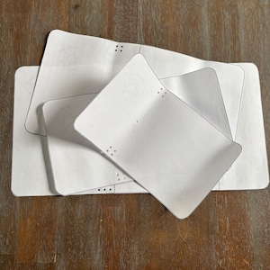 May include: Several white rectangular paper templates with rounded corners are stacked on a wooden surface. Each template has small circular cutouts and a faint circular design with text. The templates are likely for a craft or design project.