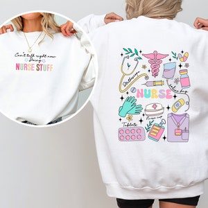 May include: White sweatshirt with a colorful graphic on the back that says "NURSE" and features medical symbols like a stethoscope, syringe, and pills. The front of the sweatshirt has the text "Can't talk right now doing NURSE STUFF".
