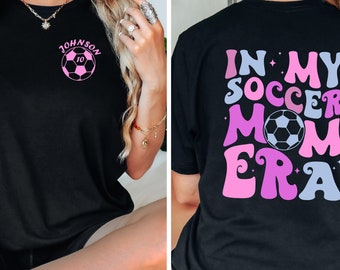 Op de afbeelding: Zwart T-shirt met een roze voetbal en de tekst "Johnson 10" op de voorkant. De achterkant van het shirt heeft de tekst "In My Soccer Mom Era" in roze, blauw en paars met een voetbal in het midden.