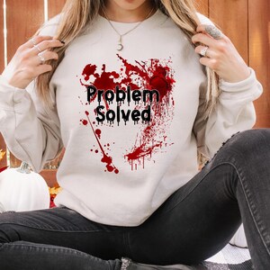 May include: A cream-colored sweatshirt with the text "Problem Solved" in black, surrounded by a splatter design in red. The sweatshirt is a long-sleeved, crew neck style. The person is wearing black jeans and black boots.
