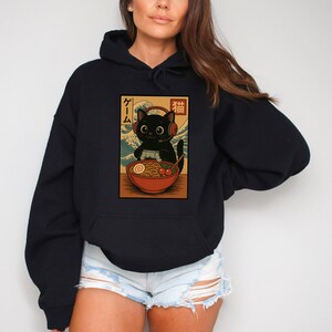 May include: Black hoodie featuring a graphic of a black cat wearing headphones, enjoying ramen. The cat is set against a backdrop of a wave pattern, with Japanese text above. The hoodie is a solid black color.