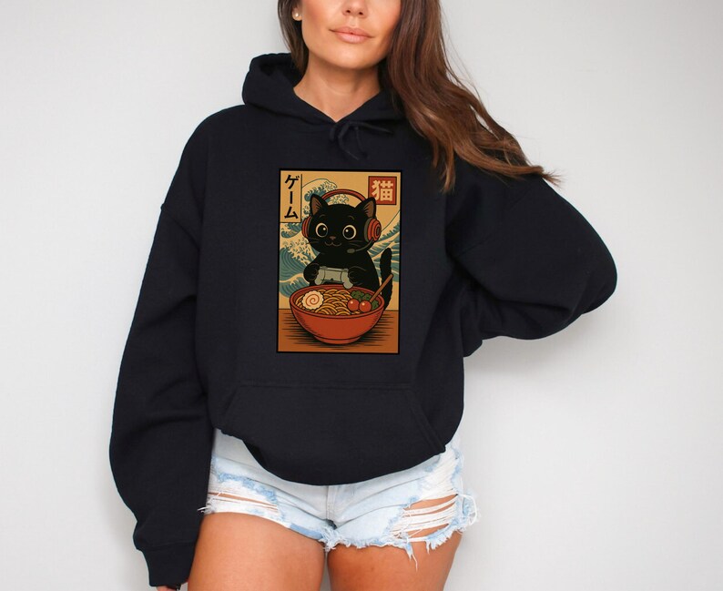 May include: Black hoodie featuring a graphic of a black cat wearing headphones, enjoying ramen. The cat is set against a backdrop of a wave pattern, with Japanese text above. The hoodie is a solid black color.