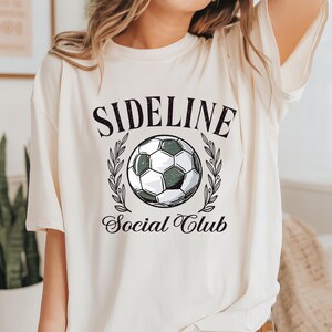 Puede incluir: Camiseta color crema con la inscripci&oacute;n "SIDELINE" sobre un gr&aacute;fico de bal&oacute;n de f&uacute;tbol, rodeado de ramas de olivo, y las palabras "Social Club" debajo. El bal&oacute;n de f&uacute;tbol es negro y blanco con un tinte verde. La camiseta tiene mangas cortas.
