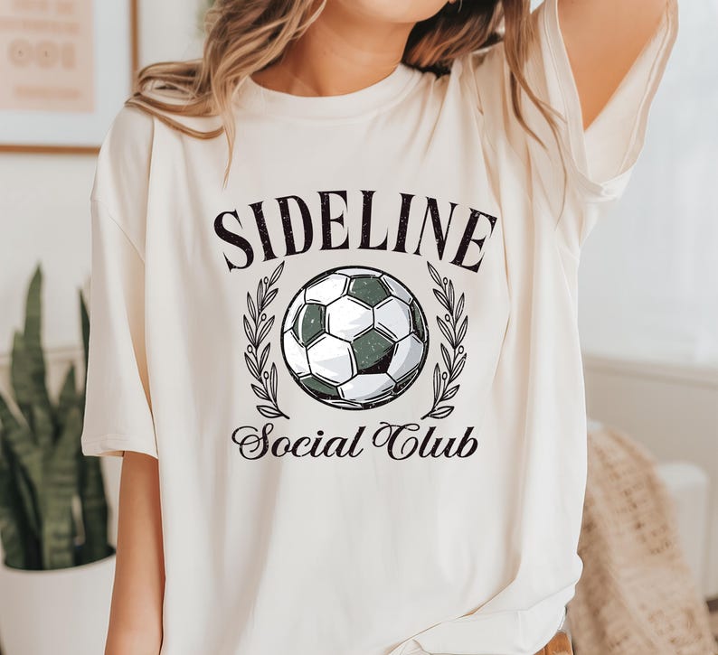 Puede incluir: Camiseta color crema con la inscripci&oacute;n "SIDELINE" sobre un gr&aacute;fico de bal&oacute;n de f&uacute;tbol, rodeado de ramas de olivo, y las palabras "Social Club" debajo. El bal&oacute;n de f&uacute;tbol es negro y blanco con un tinte verde. La camiseta tiene mangas cortas.