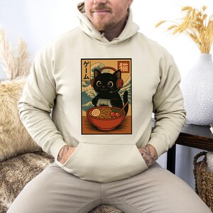 May include: Beige hoodie featuring a graphic of a black cat wearing headphones and holding a game controller, with a bowl of ramen. The graphic includes Japanese text and a wave pattern background.