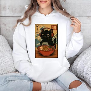 May include: White hoodie featuring a graphic of a black cat wearing headphones and playing a video game, with a bowl of ramen. The design has Japanese text and a wave pattern background.