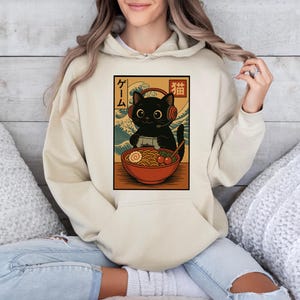 May include: A beige hoodie featuring a graphic of a black cat wearing headphones and playing a video game, with a bowl of ramen. Japanese text is on the left side of the graphic. The hoodie has a kangaroo pocket and a drawstring hood.