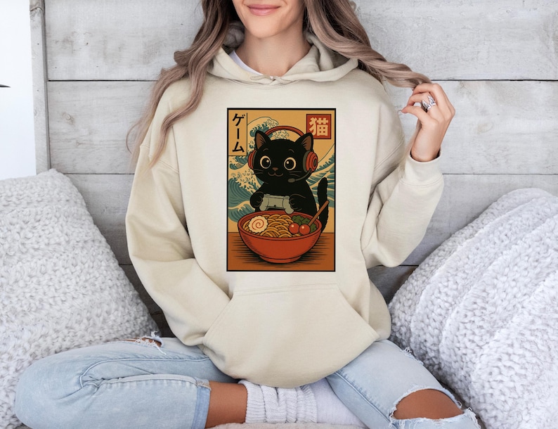 May include: A beige hoodie featuring a graphic of a black cat wearing headphones and playing a video game, with a bowl of ramen. Japanese text is on the left side of the graphic. The hoodie has a kangaroo pocket and a drawstring hood.