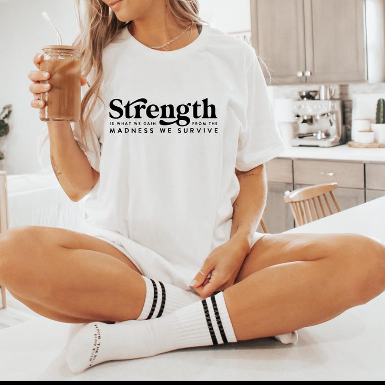 Puede incluir: Camiseta blanca con texto negro que dice "Strength is what we gain from the madness we survive".