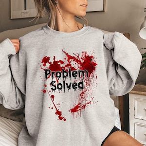 May include: A light gray sweatshirt with a graphic design. The design features the words "Problem Solved" in black, with a red splatter effect. The sweatshirt has long sleeves and a crew neck.