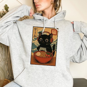 May include: A light gray hoodie featuring a graphic of a black cat wearing headphones and playing a video game. The cat is in front of a bowl of ramen. Japanese text is visible in the design.