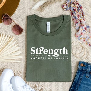 Puede incluir: Una camiseta verde con un gr&aacute;fico blanco que dice "Strength is what we gain from the madness we survive".