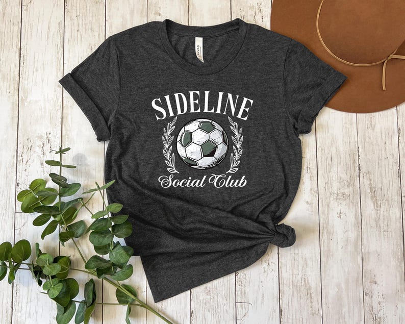 Puede incluir: Camiseta gris oscuro con las palabras "SIDELINE Social Club" y un gr&aacute;fico de bal&oacute;n de f&uacute;tbol. El dise&ntilde;o presenta un bal&oacute;n de f&uacute;tbol en el centro, rodeado por una corona de laurel. La camiseta est&aacute; anudada en la parte inferior.