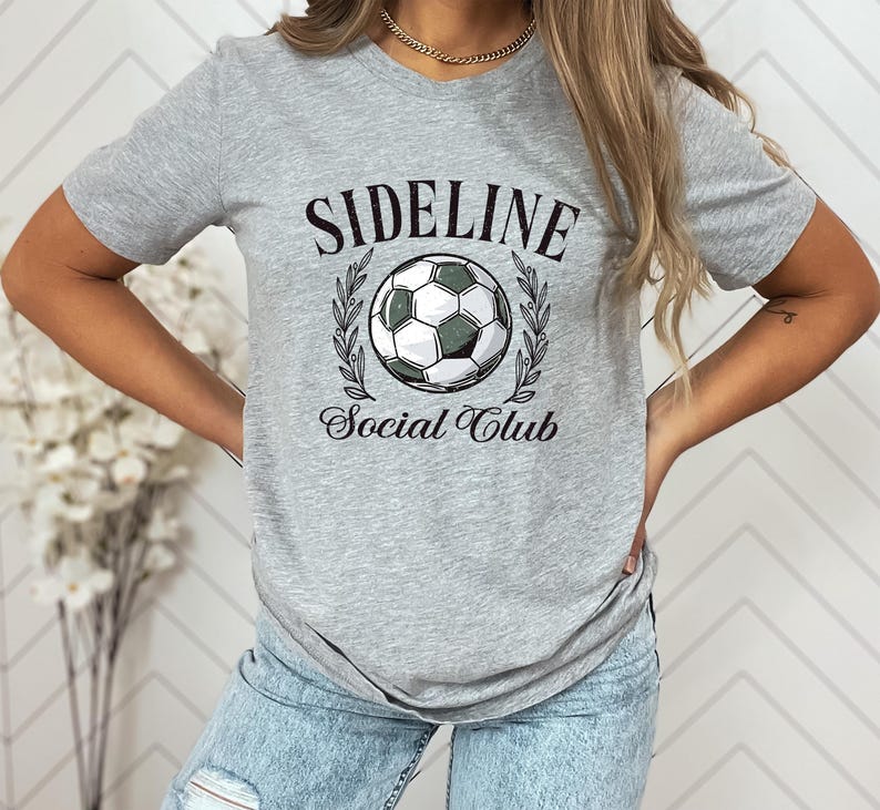 Puede incluir: Camiseta gris claro con un gr&aacute;fico de bal&oacute;n de f&uacute;tbol y las palabras "SIDELINE Social Club" en negro, con una fuente vintage. El bal&oacute;n de f&uacute;tbol est&aacute; rodeado por una corona de laurel.