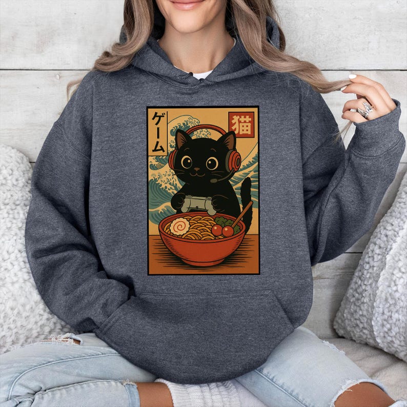 May include: Dark gray hoodie featuring a graphic of a black cat wearing headphones and playing a video game, with a bowl of ramen. The design has Japanese text and a wave pattern in the background.