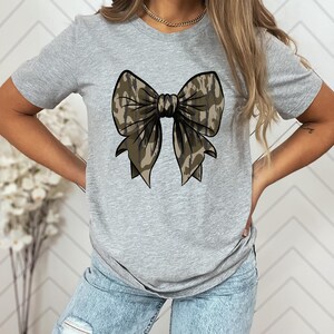 Puede incluir: Una camiseta gris con un gran lazo de camuflaje estampado en la parte delantera.